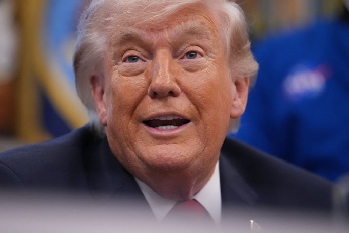A man is seen wearing a suit, with a partially lifted flap from the camera angle. The background is out of focus, with an indistinct emblem on what might be a flag or other symbols. The overall setting appears to be indoors, possibly at a formal event or press conference.