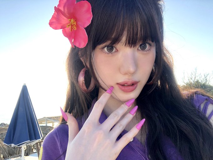 A close-up of a woman with dark hair, pink nails, and a pink hibiscus flower in her hair, posing against a bright, sunny background.