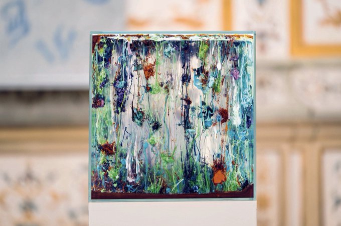 The image features a painting with blue, green, red, and white colors that appears to be abstract or surrealistic. The artwork is displayed prominently against a blurred background, which could be an art gallery or a room filled with other paintings. The painting's composition and use of color evoke a sense of creativity and imagination.