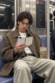 A man in a brown blazer and khaki pants sits on a train seat, looking down at his phone while holding a drink, suggesting he's focused on his screen rather than his surroundings.