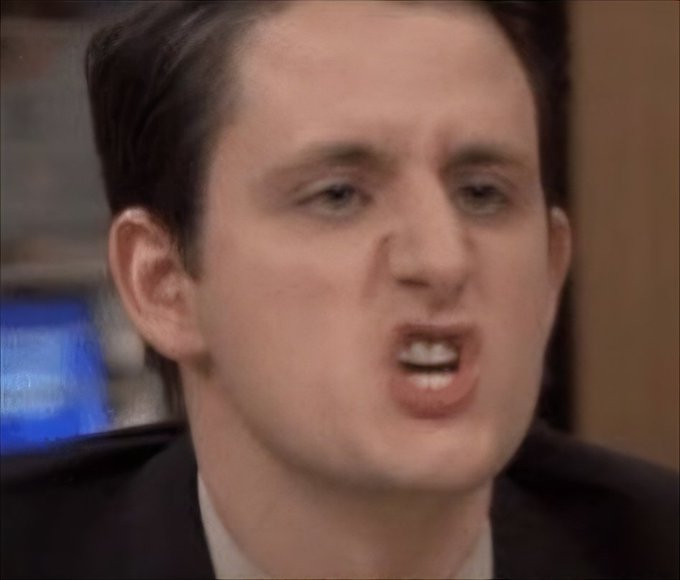 Close-up of a man with dark hair making an intense, wide-eyed facial expression with his mouth open, wearing a dark suit jacket and light shirt.