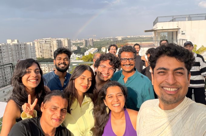 A group selfie of several smiling people on a rooftop with a rainbow visible in the sky and a cityscape in the background.