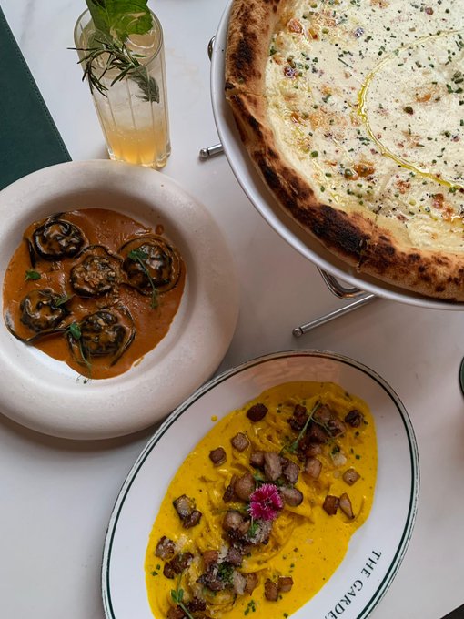 The image features an inviting spread of gourmet dishes, including a wood-fired pizza with a bubbly, herb-garnished crust, a creamy orange-hued pasta with rolled eggplant slices, and a vibrant yellow dish topped with diced potatoes and garnished with a pink flower, all elegantly presented on white plates—one reading "THE GARDEN." This beautifully plated meal reflects the post's theme of taking a well-deserved break to recharge with good food while continuing the creative journey with Ritual.