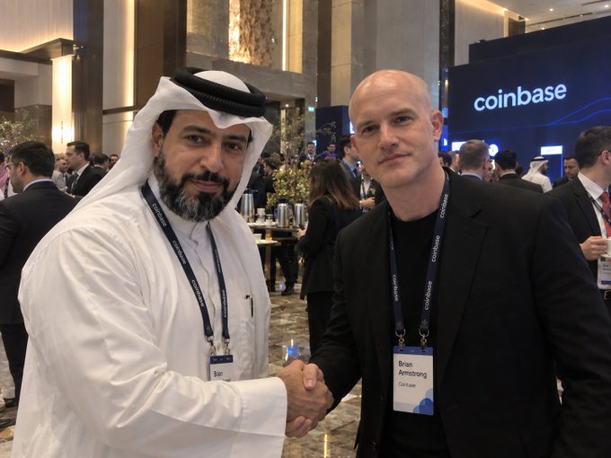 Two men shake hands at a Coinbase event, with name tags identifying one as 'Brian Armstrong' from Coinbase.