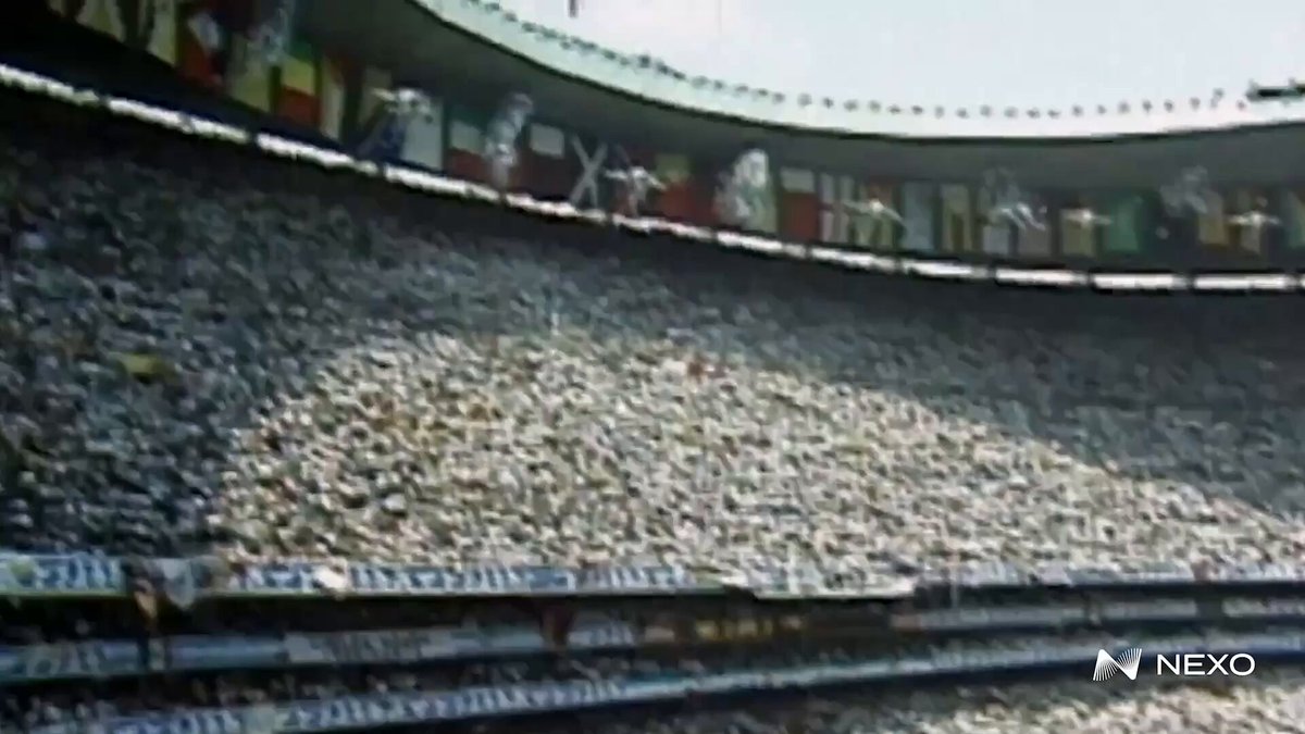 The image shows a large stadium filled with people watching a soccer game. The crowd appears to be diverse, consisting of individuals from various countries. The stadium has multiple levels, indicating that it can accommodate a significant number of spectators. The perspective of the photo is taken from below, looking up at the crowd and the stadium's architecture.
