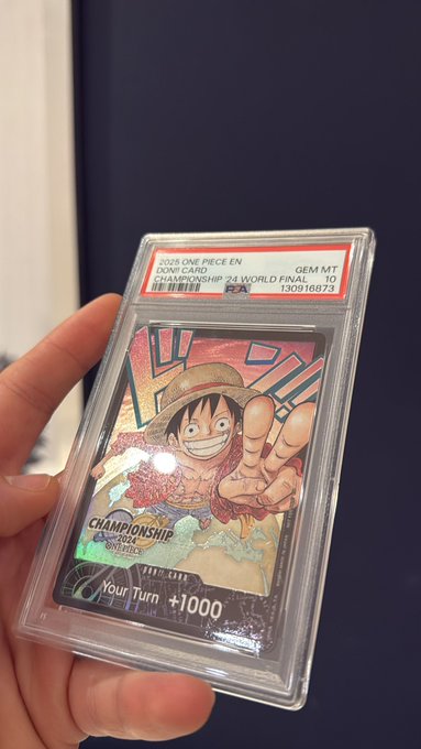 A hand holding a PSA-graded PSA 10 GEM MT trading card of Monkey D. Luffy from the "2025 ONE PIECE TCG EN CHAMPIONSHIP 24 WORLD FINAL" featuring the Championship 24 logo and "Your Turn +1000" text.