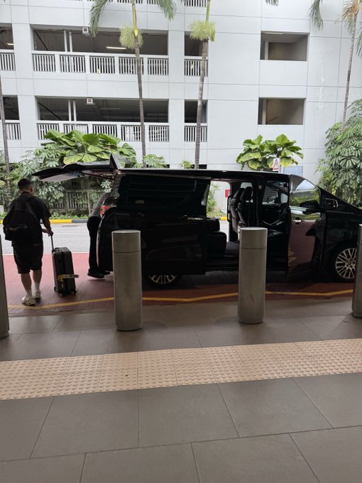 A man with a backpack and rolling suitcase approaches a sleek black VIP van at an airport drop-off zone, where staff assist with luggage before his ride. The image captures the premium airport transfer experience being offered as part of the TOKEN2049 VIP promotion by Kast.