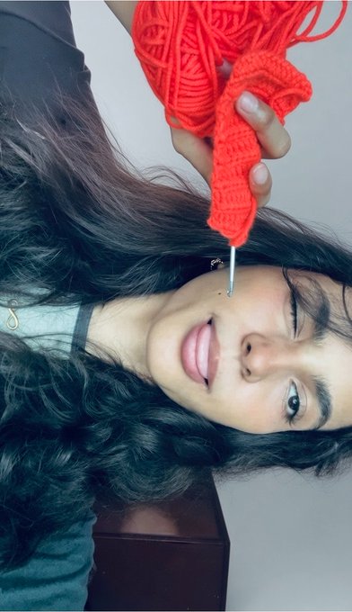 A young woman with long dark hair lying down while holding red crocheted items near her face.