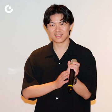 A smiling young man with dark hair, wearing a black short-sleeved button-up shirt, holds a microphone with both hands as if speaking at an event, with a "G" logo visible in the upper left corner of the image, likely representing Gate Charity's gathering of VIP guests and industry partners.