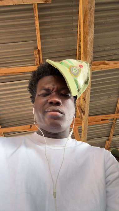 A young man wearing a light green John Deere cap and white shirt makes a playful, tired expression while taking a selfie under a wooden-framed metal roof, capturing the exhausted but relieved mood of nearly finishing his "11/12" task.