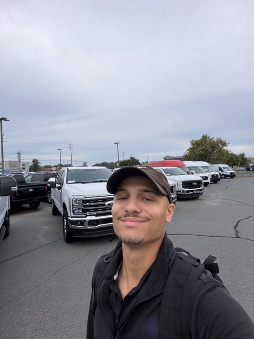 A selfie of a smiling man with a mustache wearing a black cap and jacket with a backpack strap, taken in a car dealership lot with several Ford trucks visible behind him.