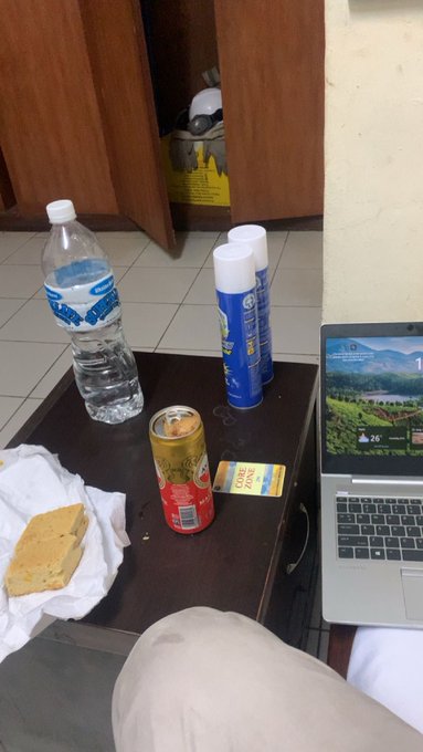 A casual scene on a wooden table with a bottle of water, two aerosol cans, a soda can, a tissue with food, and an open laptop showing a wallpaper, with a person's leg visible in the foreground.