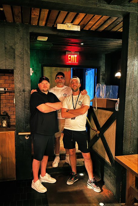Three men pose together in a dimly lit interior beneath an illuminated red EXIT sign, with one man in the center wearing a white t-shirt and glasses.
