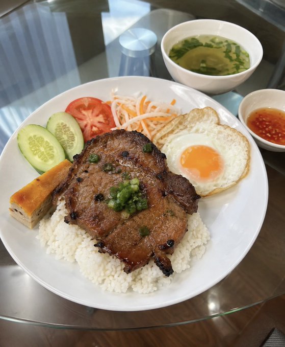 A Vietnamese-style rice plate featuring a grilled pork chop on rice, accompanied by a fried egg, cucumber slices, tomato, pickled carrots, and a bowl of broth with dipping sauce.
