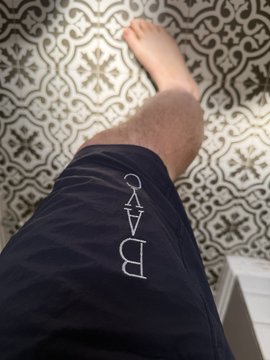 A photograph looking down at a person's leg wearing dark blue swim trunks with the word 'CAVB' embroidered on the side, with a patterned tile floor visible below.