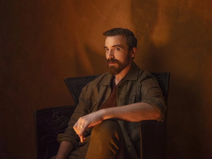 A bearded man with slicked-back hair sits in a dark leather armchair against a warm, amber-lit backdrop, gazing thoughtfully off-camera. The intimate portrait complements the birthday tribute, presenting Sam Spratt—the visionary creator of LUCI—in a contemplative and artistic light befitting his status as a defining artist of our time.