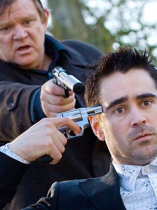 A tense scene showing a man in a suit having a silver revolver pointed directly at his temple by another man standing behind him in a dark jacket.