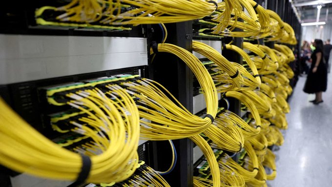 Rows of bright yellow fiber optic cables are meticulously routed through server racks in a data center, illustrating the massive physical infrastructure required to support the GPU compute power that the post warns is outpacing available electricity capacity. The image visually underscores the growing tension between expanding AI hardware demand and the real-world constraints of power and buildout that the accompanying text highlights.