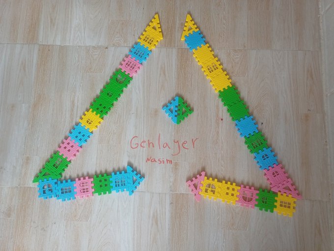 A child's colorful block creation arranged on a wooden floor to form a triangular house-like structure with the handwritten text "Geblager nasim" inside.