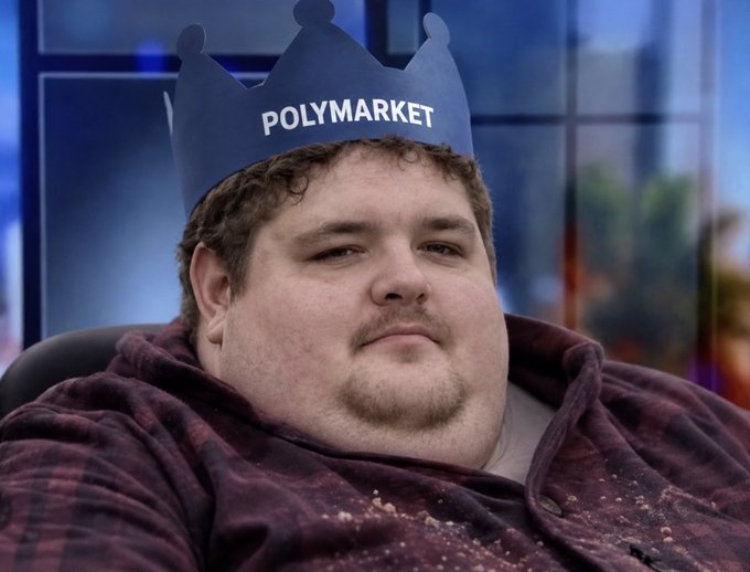 A young man with curly hair wearing a red plaid shirt and a blue paper crown labeled "POLYMARKET" on his head, sitting in a chair with a neutral expression.
