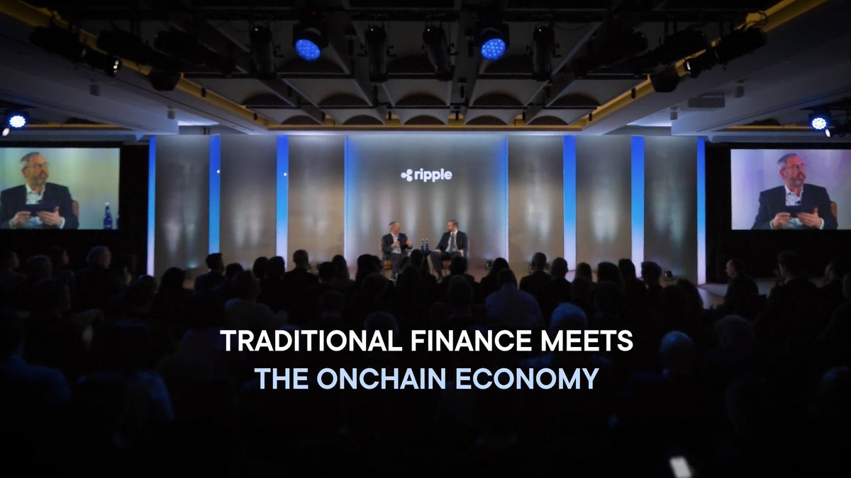 A conference event with a stage displaying the Ripple logo, showing two speakers seated before an audience, with large screens flanking them and text reading 'Traditional Finance Meets the Onchain Economy.'