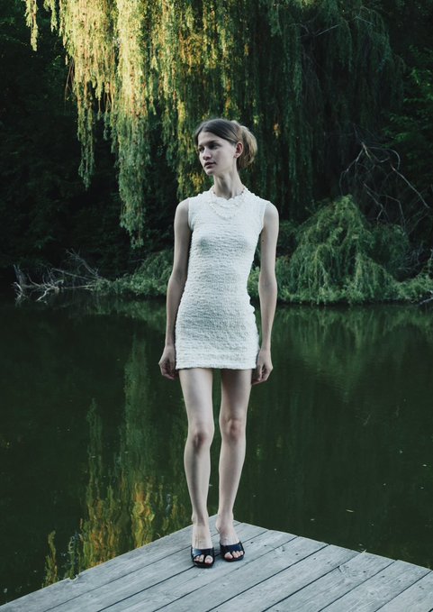 A female model in a sleeveless, textured white mini-dress stands barefoot in black sandals on a wooden dock next to a dark green body of water, surrounded by overhanging trees.