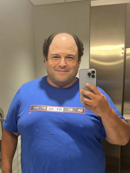 A balding man takes a mirror selfie with a silver smartphone while wearing a blue t-shirt in what appears to be a public restroom with metal stalls.