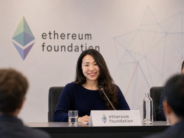 The image shows a woman sitting at a table with two other people behind her. They are all gathered around a conference table, which has a sign that says "Ethereum Foundation". The woman appears to be smiling as she looks directly at the camera. In the background, there is another person who seems to be looking away from the camera. The image also includes text about personnel changes within the organization and mentions two individuals leaving their positions: Hsiao-Wei Wang and Tomasz Stańczak.