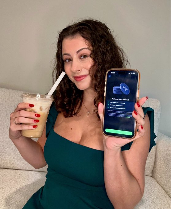 The image features a woman sitting on a couch holding up her phone with a drink in front of her. The woman has red nail polish on her nails and is wearing a green dress. She appears to be posing for a photo, possibly sharing her experience or promoting an exchange related to passive income.