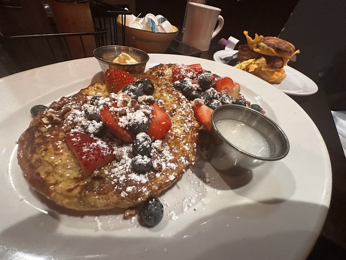 A restaurant breakfast plate features pancakes topped with fresh strawberries, blueberries, and powdered sugar, accompanied by a small pitcher of syrup, with a side dish visible in the background.