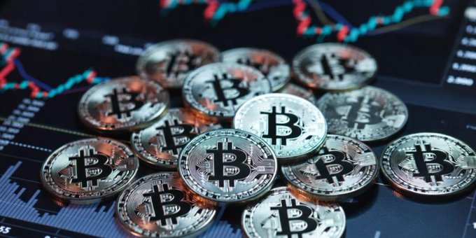 The image shows a collection of Bitcoin coins scattered across a black and white background. A blue text overlay suggests that these Bitcoins may be from Standard Chartered Bank, though there's no actual bank logo visible in the image. The text also mentions "floor" and "USD 100,000". These Bitcoins might have been affected by various market conditions, such as inflation or fluctuations in value due to economic factors like interest rates or central bank policies.