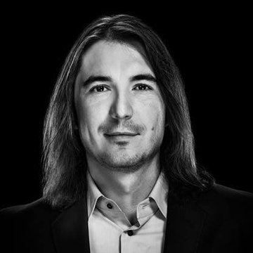 A black-and-white headshot portrait of a man with long hair, wearing a suit jacket and light button-up shirt, looking directly at the camera.
