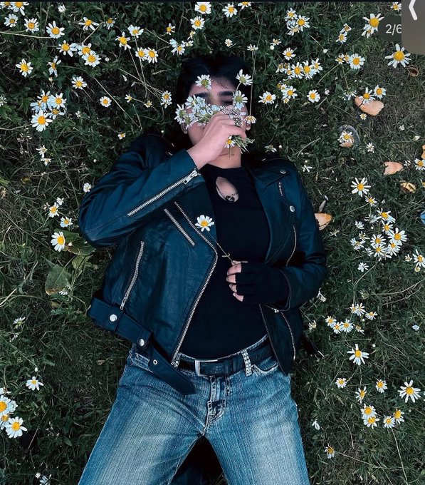 A person lies on grass covered in white daisies, holding flowers over their face, wearing a black leather jacket and blue jeans in a 2/6 Instagram carousel.