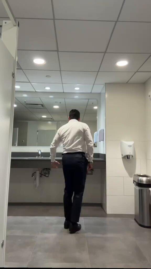 The image shows a man standing in front of a sink, possibly washing his hands or face. The bathroom has white walls and gray floor tiles, giving it a clean and modern appearance. A mirror can be seen above the sink, reflecting the room's interior. There is also a trashcan present in the scene.