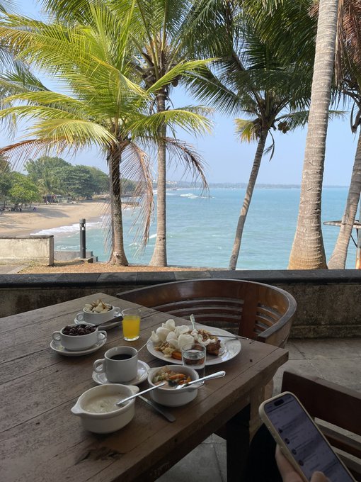 A breakfast spread with coffee, juice, and various dishes sits on a wooden table overlooking a tropical beach lined with palm trees, with someone scrolling on their phone nearby—capturing a moment of leisure before returning to "degen" activities.