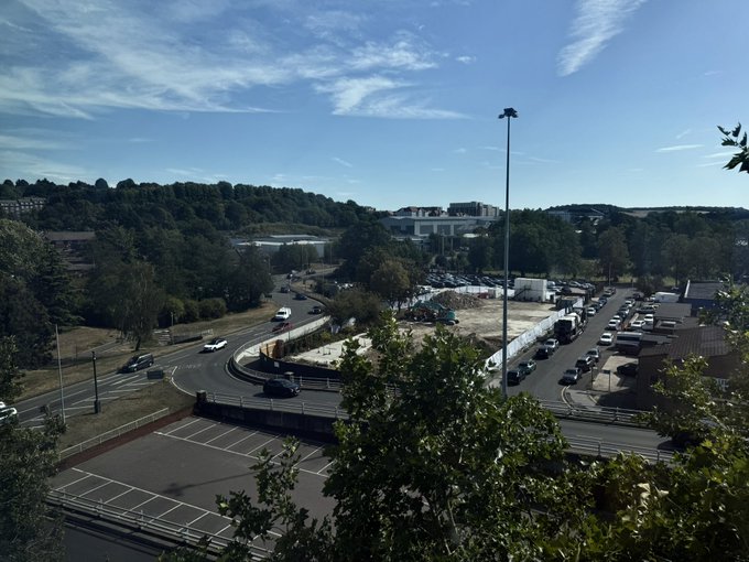 A sunny morning view from above shows a curving road, parking lots, and buildings surrounded by green hills under a bright blue sky, capturing the everyday scenic outlook the poster wakes up to.