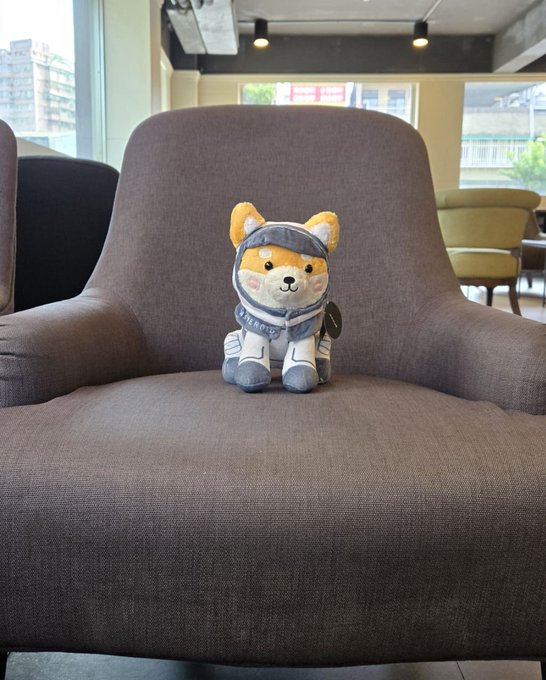 A Shiba Inu plush toy dressed in a blue and white outfit sits on a gray armchair in a modern indoor setting with large windows.