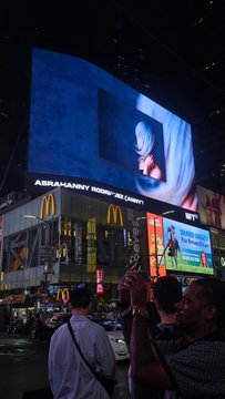 A nighttime photograph of Times Square in New York City shows large illuminated billboards, including advertisements for McDonald's and a large screen displaying 'ASIAN AMERICAN SOCIETY OF AMERI', with crowds of people and a man taking a photo in the foreground.