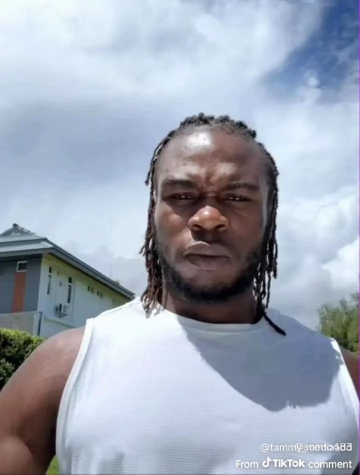 A man with braided hair wearing a white tank top stands outside with a building and cloudy sky behind him, with a TikTok comment watermark visible.