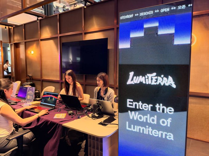 A booth at an event featuring women working on laptops behind a table, with a large illuminated display reading 'Lumiterra' and 'Enter the World of Lumiterra'.