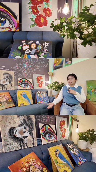 A woman sits on a blue couch in an art studio displaying various colorful paintings including eyes, flowers, a panda, and parrots.