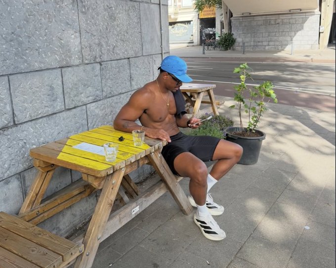 A shirtless man in a blue cap and black shorts sits on a sunny sidewalk picnic bench checking his phone, captioned "GM CT ☀️ Happy Sunday everyone." The relaxed weekend vibe of the image matches the casual "Who's active?" engagement post encouraging the NomismaNetwork community to start their Sunday off together.
