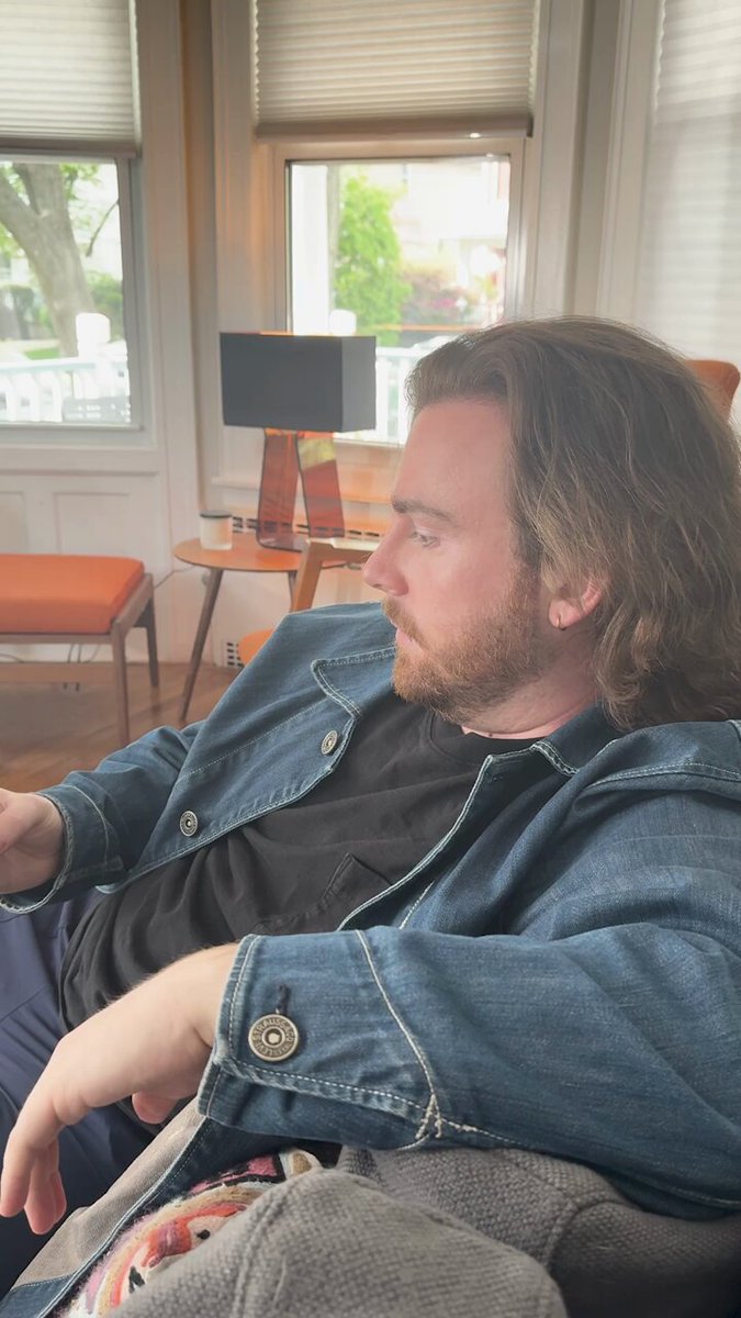 The image shows a man sitting comfortably on a couch with his arm resting on another person's shoulder, possibly indicating a close relationship between them. The background features a window that allows natural light into the room, creating a cozy atmosphere. There are also two chairs in the scene - one near the man and the other further away from him.