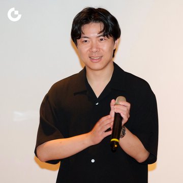 A smiling Asian man in a black short-sleeved shirt stands holding a microphone, appearing to be speaking at an event.