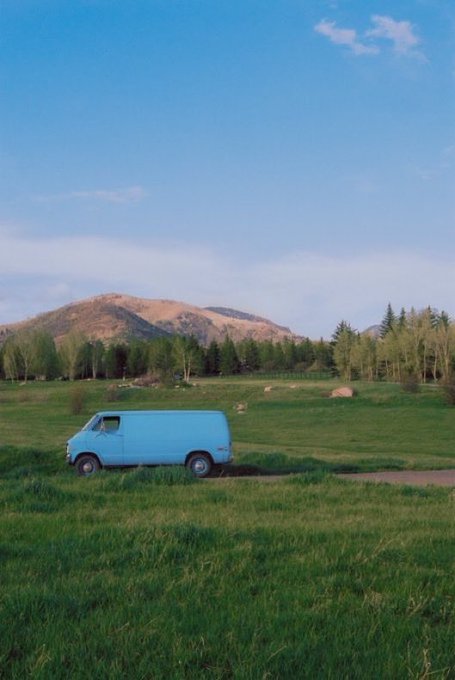 A blue vintage van parked in a grassy field with a mountain and trees in the background under a clear sky.