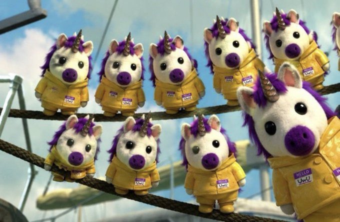 Several plush toys resembling unicorn pigs wearing yellow jackets with name tags sit on ropes against a backdrop of a blue sky and what appears to be a ship.