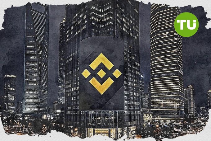 A black-and-white cityscape of illuminated skyscrapers featuring the Binance logo overlaid in the center, with a green 'TU' (The Defiant) badge in the top right corner.