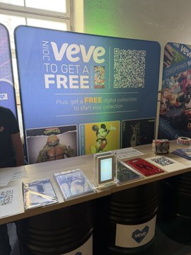 The image features a table with various items displayed on it, including a sign that says "VeVe just announced integrations with both Telegram AND Roblox. That's 1B+ and 380M+ monthly active users respectively." The table also has several books and CDs placed on top of it. In the background, there is a person standing near the table.