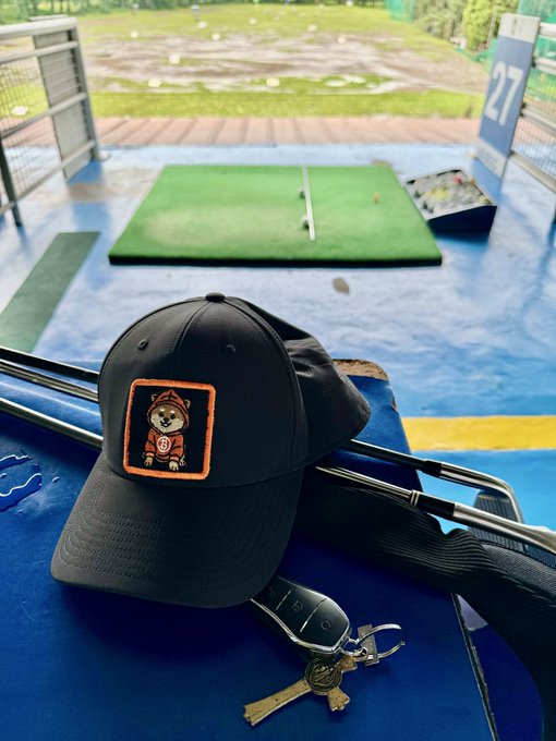 A black baseball cap with an orange fox patch rests on golf clubs at a driving range, with keys lying in the foreground and a practice mat and field visible in the background.