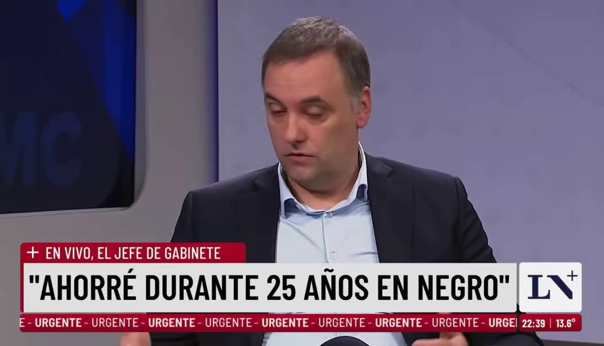 The image shows a man sitting at a desk with a TV screen behind him, displaying a news segment or broadcast. The man appears to be wearing a suit and tie, suggesting that he might be a professional or an important figure. The TV screen is in Spanish, which indicates that the content being displayed is likely related to the country of origin for the man sitting at the desk.